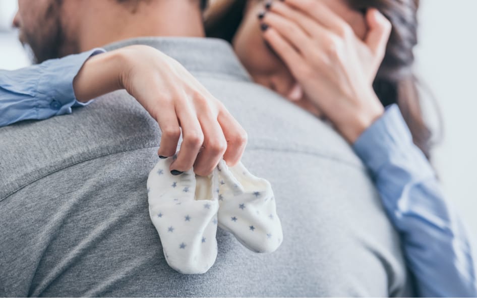 Sad infertile couple hugging, woman holding baby shoes symbolizing infertility struggle